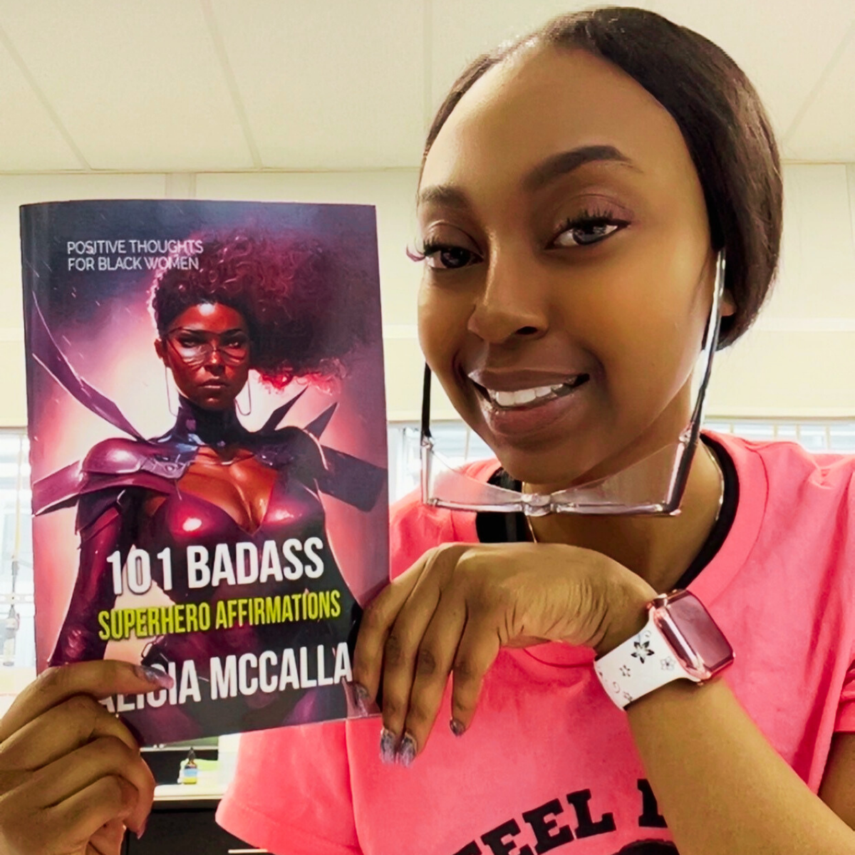Woman in a pink superhero shirt smiling and holding Alicia McCalla’s 101 Badass Superhero Affirmations book with the cover facing forward.